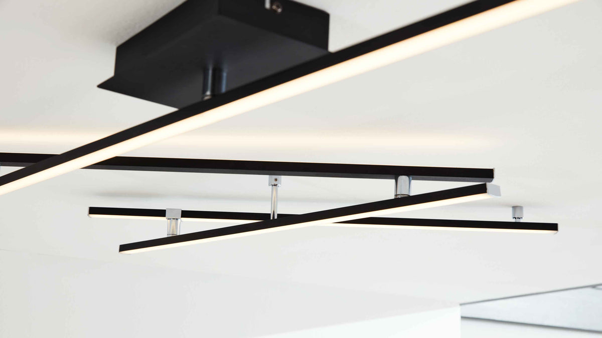 Deckenleuchte mit mehreren sich kreuzenden schwarzen LED-Linienstäben, an silbernen Halterungen an weißer Decke montiert.