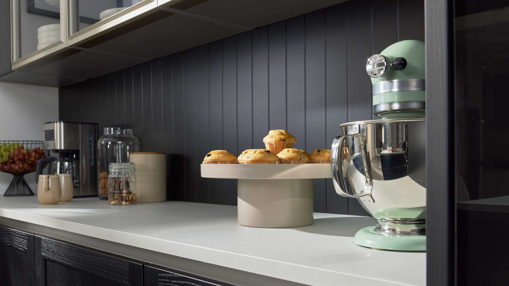 Beiger Tortenständer mit Muffins auf Arbeitsplatte, rechts mintgrüner Küchenmixer, links Kaffeemaschine und Vorratsgläser.