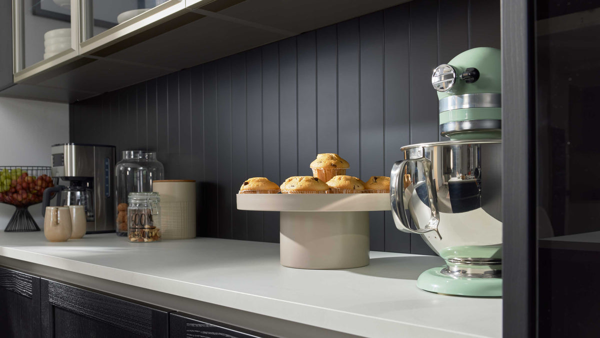 Beiger Tortenständer mit Muffins auf Arbeitsplatte, rechts mintgrüner Küchenmixer, links Kaffeemaschine und Vorratsgläser.