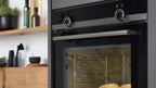 Schwarzer Einbaubackofen mit Edelstahlgriff, zwei Drehreglern und Sichtfenster, innen Backbleche mit Brötchen; links Holzplatte mit Flaschen.