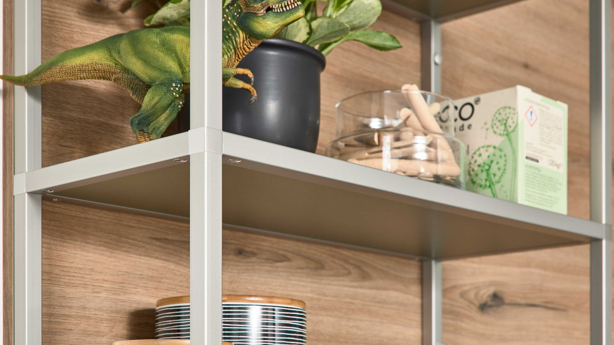 Offenes Regal mit silbernem Metallrahmen und Holzrückwand, darauf Blumentopf, grünes Dinosaurierspielzeug und Glas mit Holzklammern.