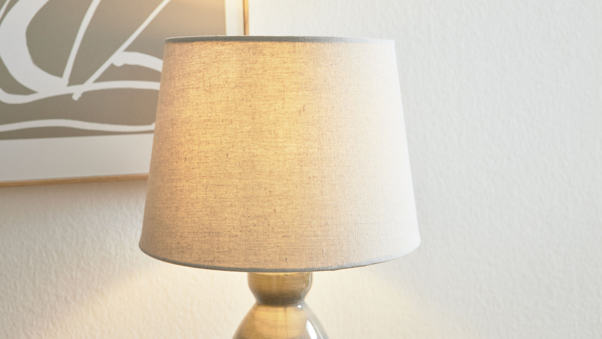 Tischlampe mit hellem, leicht strukturiertem Stoffschirm und glasiertem Keramikfuß vor weißer Wand und Bildrand.