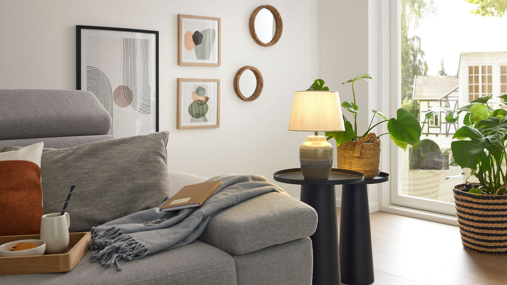 Graues Polstersofa mit Kissen, Decke und Notizbuch, daneben zwei schwarze Beistelltische mit Lampe und Topfpflanze am Fenster.