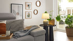 Graues Polstersofa mit Kissen, Decke und Notizbuch, daneben zwei schwarze Beistelltische mit Lampe und Topfpflanze am Fenster.