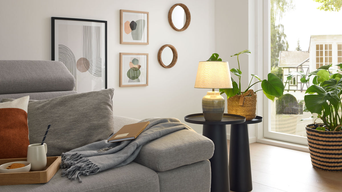 Graues Polstersofa mit Kissen, Decke und Notizbuch, daneben zwei schwarze Beistelltische mit Lampe und Topfpflanze am Fenster.