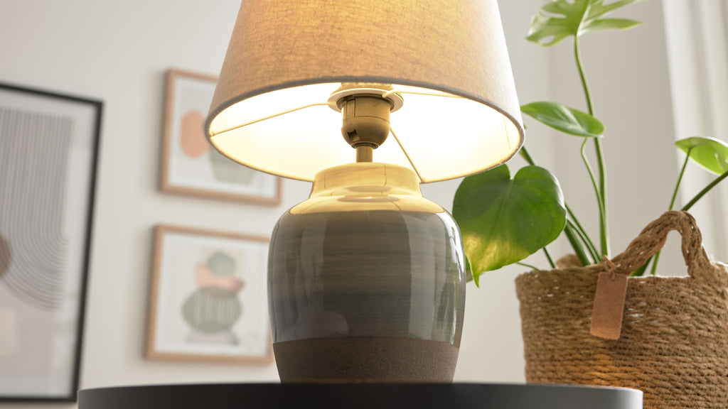 Keramik-Tischlampe mit beigem Stoffschirm auf schwarzem Tisch, rechts eine Pflanze im Korb und unscharfe Bilder.