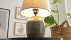 Keramik-Tischlampe mit beigem Stoffschirm auf schwarzem Tisch, rechts eine Pflanze im Korb und unscharfe Bilder.