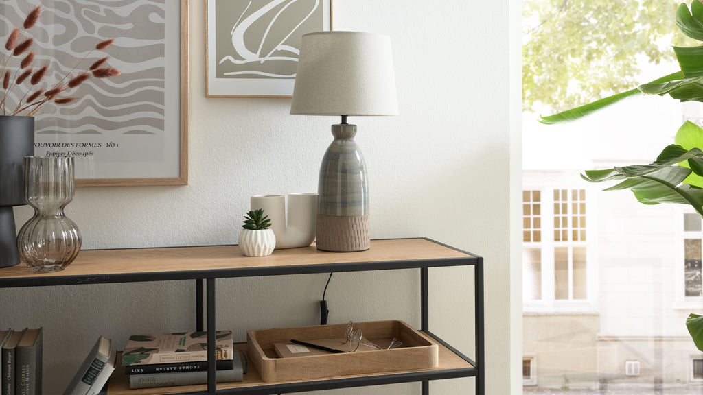 Tischlampe mit beigefarbenem Stoffschirm auf schmalem Holz-Metall-Regal, daneben kleine Sukkulente, Vasen, Bücher und Tablett.