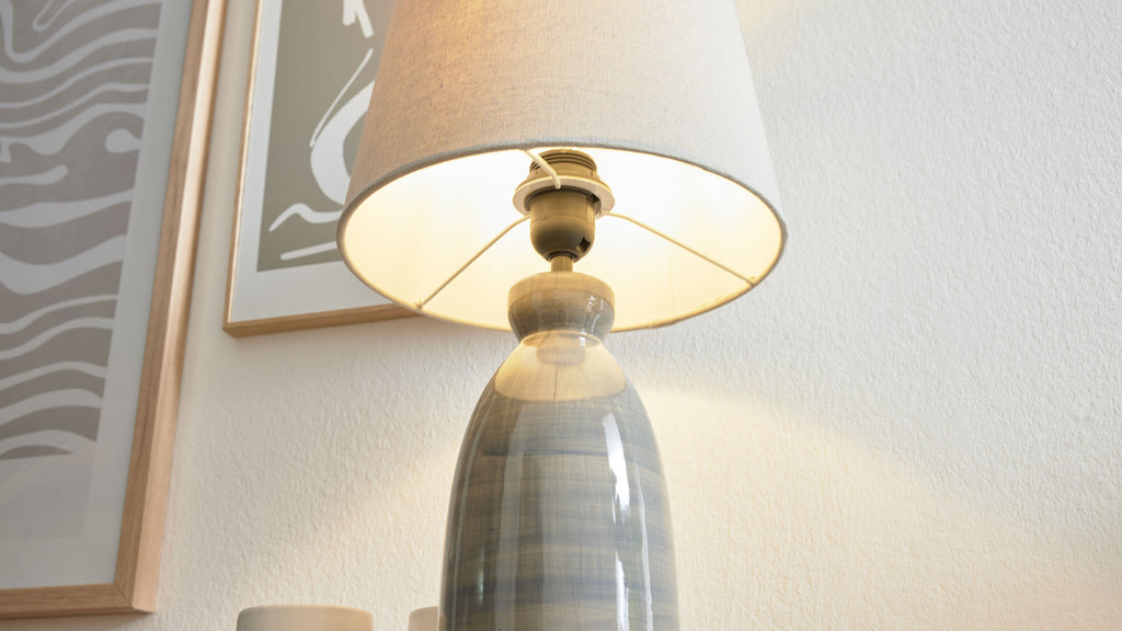 Tischlampe mit glasiertem blau‑grau gestreiftem Keramikfuß und beigem Stoffschirm, links zwei gerahmte Bilder an der Wand.
