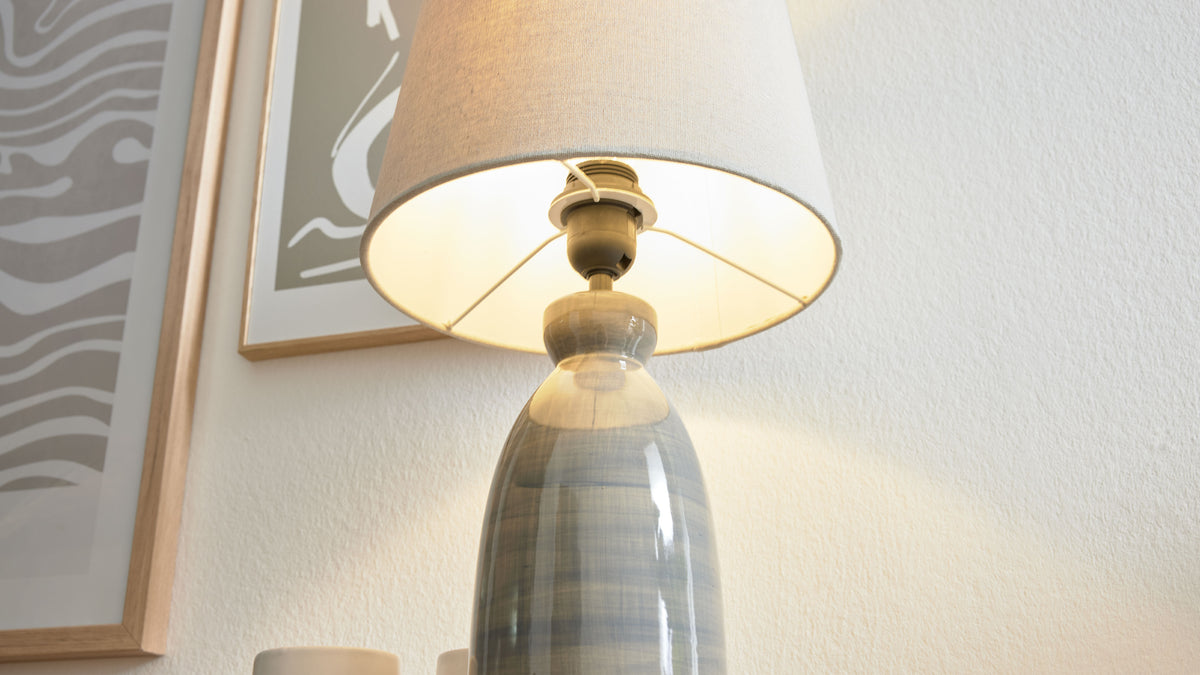 Tischlampe mit glasiertem blau‑grau gestreiftem Keramikfuß und beigem Stoffschirm, links zwei gerahmte Bilder an der Wand.