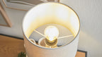 Tischlampe mit beige Stoffschirm und hell leuchtender Glühbirne, sichtbare Metallstreben, auf Holzplatte neben Wand und Rahmen.