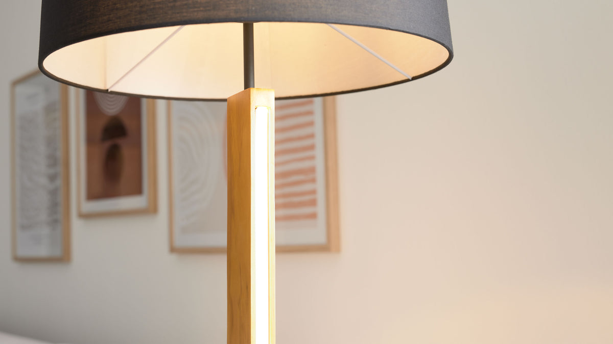 Lampe mit großem dunkelgrauem Stoffschirm und einem hellen vertikalen Leuchtstreifen im Holzschaft, vor unscharfen gerahmten Bildern.