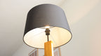 Stehende Lampe mit großem dunkelgrauem Stoffschirm, sichtbarer Leuchtbirne, Metallgestänge und rechteckigem Holzfuß, im Raum.