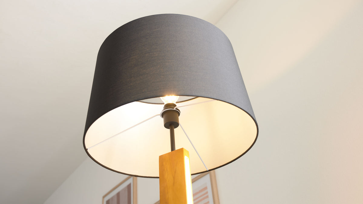 Stehende Lampe mit großem dunkelgrauem Stoffschirm, sichtbarer Leuchtbirne, Metallgestänge und rechteckigem Holzfuß, im Raum.
