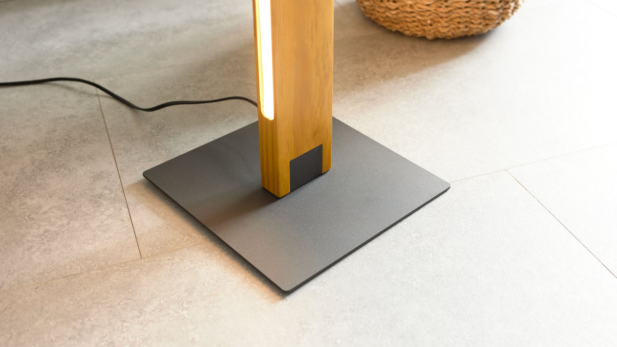 Schwarzer quadratischer Lampenfuß aus Metall mit senkrechtem Holzständer und eingebauter Leuchtleiste, Kabel und helle Fliesen sichtbar.