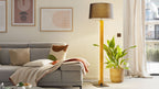 Hohe Stehlampe mit schwarzem Schirm und hellem Holzfuß neben grauem Ecksofa mit Decke und Kissen sowie Korb mit Grünpflanze.