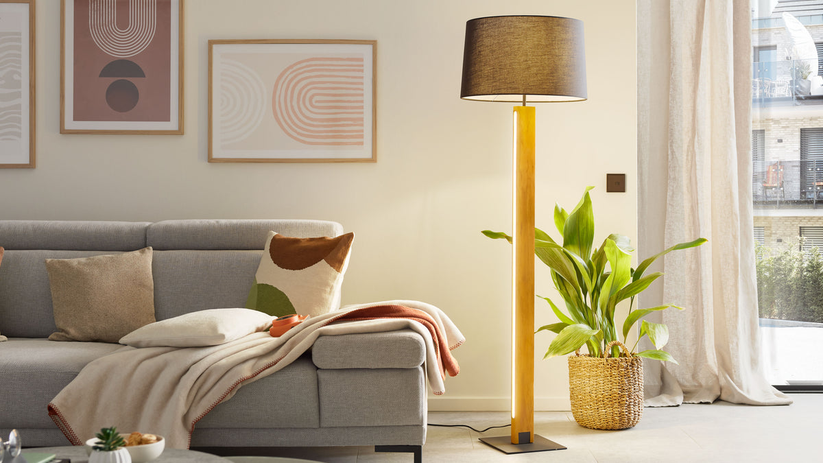 Hohe Stehlampe mit schwarzem Schirm und hellem Holzfuß neben grauem Ecksofa mit Decke und Kissen sowie Korb mit Grünpflanze.