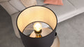 Schwarze Stehlampe mit beleuchtetem Glühbirnenkorpus in hellem Innenstoff, im Hintergrund hellgraues Sofa mit zwei Kissen.