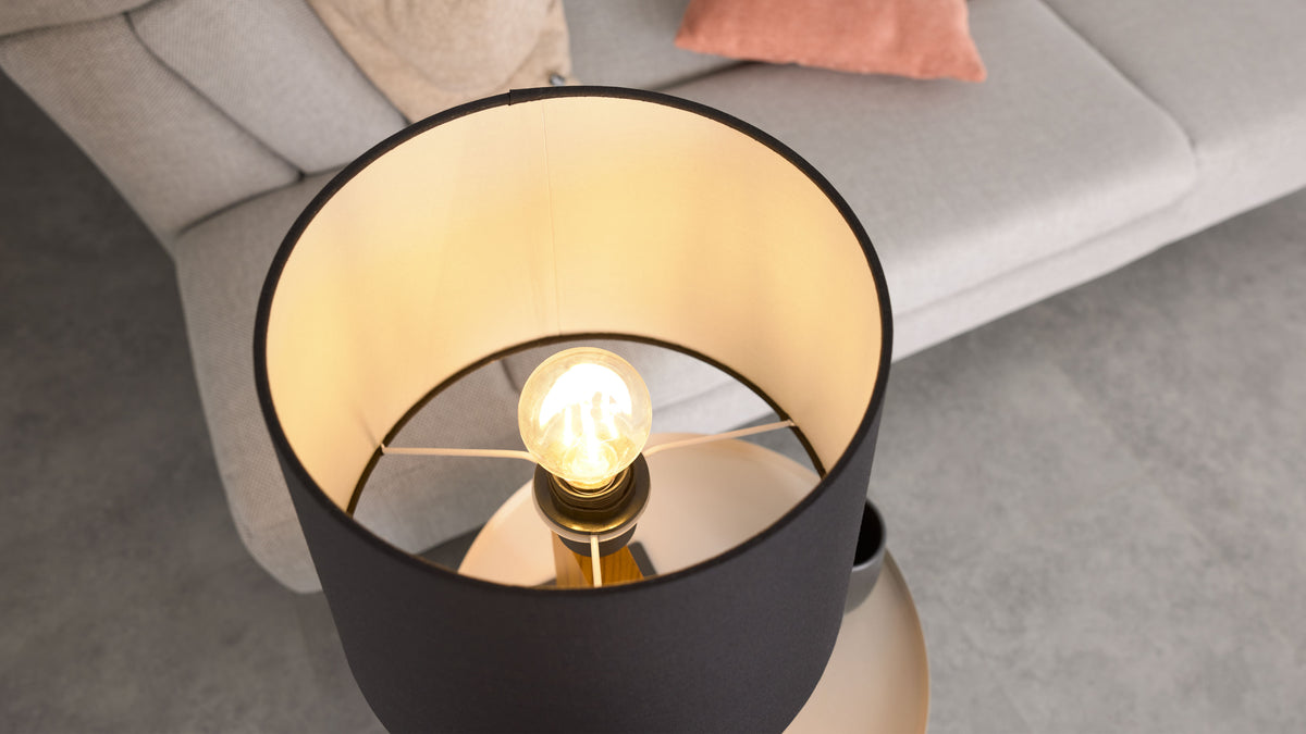 Schwarze Stehlampe mit beleuchtetem Glühbirnenkorpus in hellem Innenstoff, im Hintergrund hellgraues Sofa mit zwei Kissen.