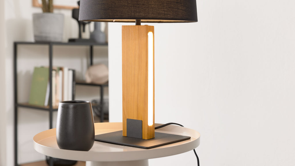 Tischlampe mit rechteckigem Holzfuß, schmalem LED-Streifen und schwarzem Schirm auf rundem Tisch; links schwarzer Becher, im Hintergrund Regal.