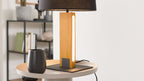 Tischlampe mit rechteckigem Holzfuß, schmalem LED-Streifen und schwarzem Schirm auf rundem Tisch; links schwarzer Becher, im Hintergrund Regal.