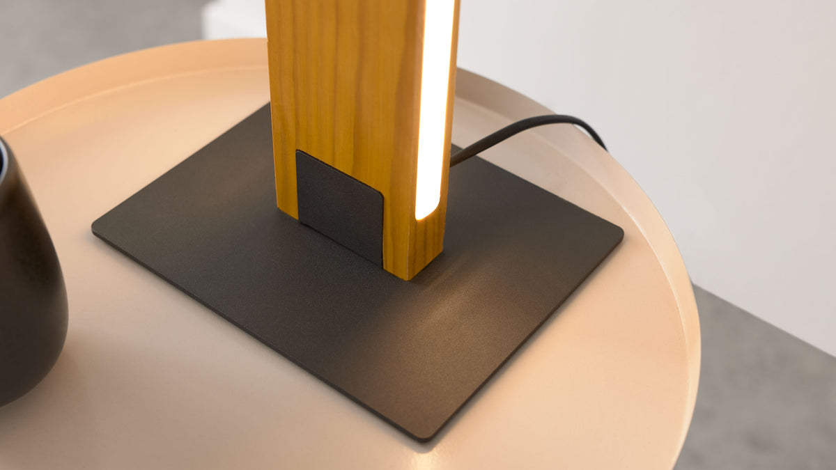 Holz-Leuchtkörper einer Tischlampe mit vertikalem Streifenlicht auf schwarzer Metallplatte, auf rundem hellrosa Tisch mit schwarzem Kabel.