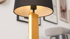 Tischlampe mit schwarzem Stoffschirm und warmem Innenlicht, Holzfuß mit vertikalem Leuchtstreifen, im Hintergrund gerahmte Bilder.