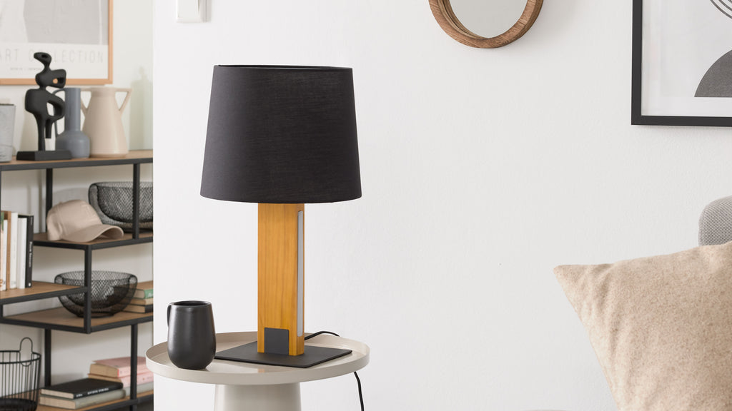 Tischlampe mit schwarzem Stoffschirm und Holzständer auf rundem Beistelltisch, daneben kleine schwarze Vase und Regal im Hintergrund.