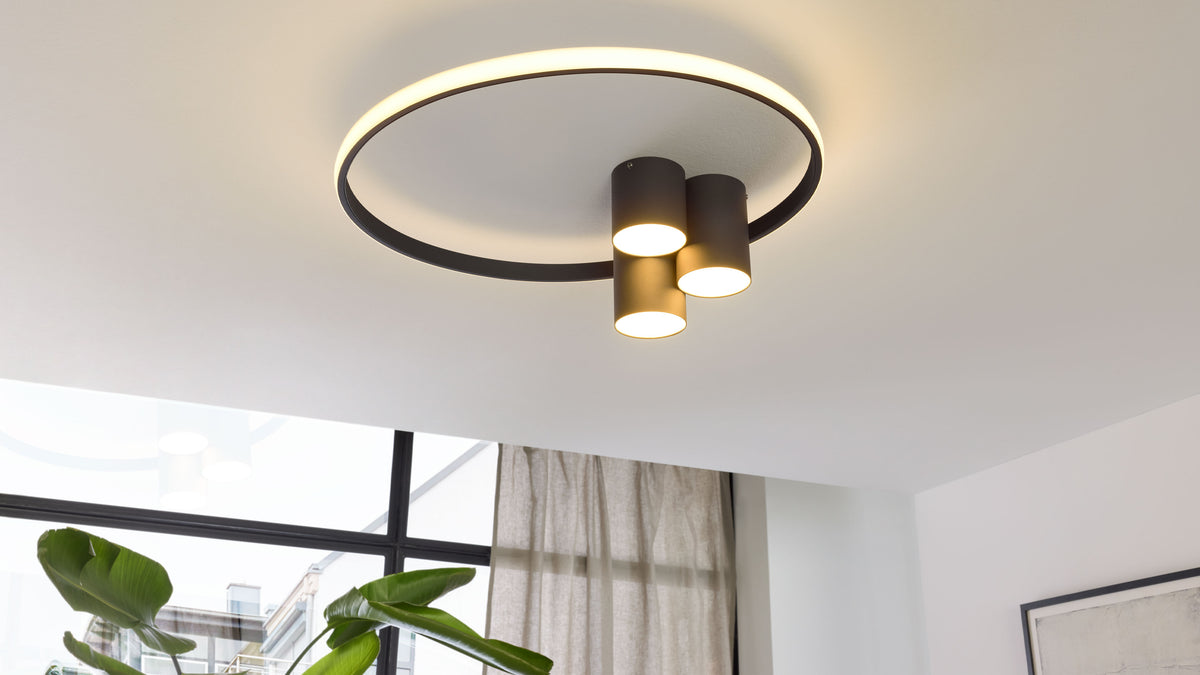 Deckeuchte mit einem großen leuchtenden Ring und drei schwarzen zylindrischen Downlights, die warmes Licht nach unten werfen.