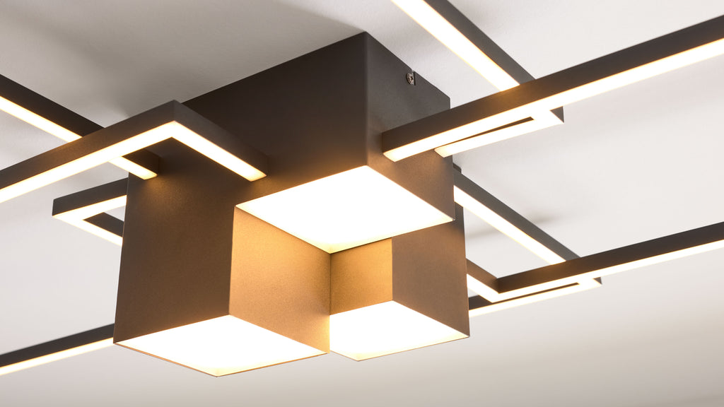 Deckeuchte mit matt-dunklem Metallgestell, gekreuzten beleuchteten Stabprofilen und drei leuchtenden quadratischen Schirmen.