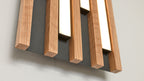 Wandmontiertes Paneel mit mehreren parallelen, quadratischen Holzleisten vor einer dunklen Rückwand und schmalen hellen Leuchtstreifen.