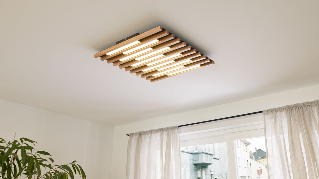 Deckenleuchte mit parallel angeordneten hell leuchtenden Holzlamellen in Zimmermitte, dazu Gardinen vor einem Fenster und eine Zimmerpflanze links.