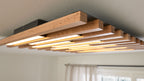 Rechteckige Deckenleuchte mit horizontalen Holzlamellen und integrierten warmen LED-Streifen, schwarzer Montagekasten sichtbar.