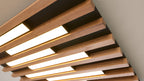Deckeninstallation aus parallelen Holzlamellen mit eingelassenen rechteckigen, warm leuchtenden LED-Paneelen in schwarzem Rahmen.