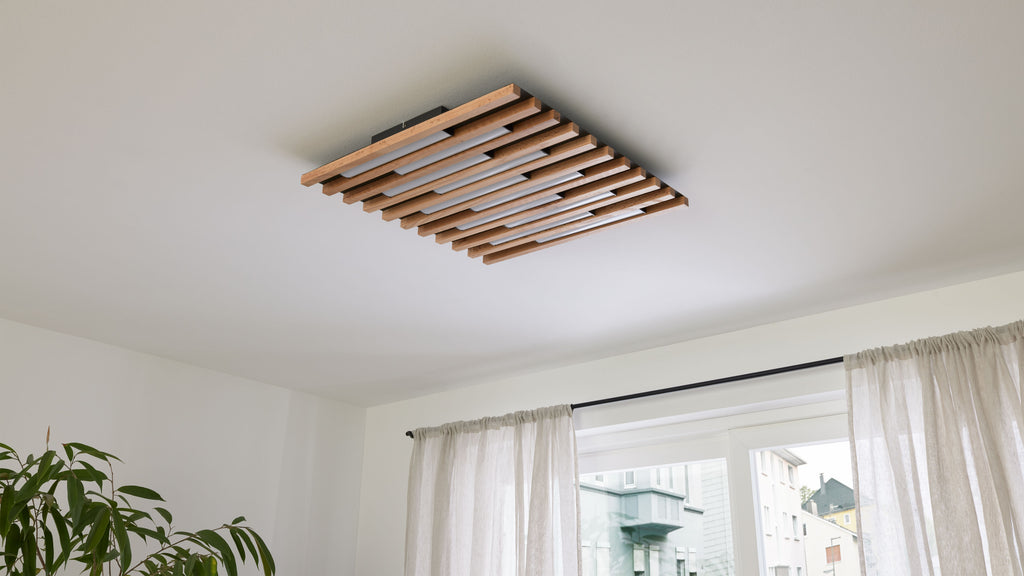 Quadratische Deckenleuchte aus parallelen Holzlatten mit weißen Leuchten, an weißer Decke über Fenster mit leichten Vorhängen und Pflanze.