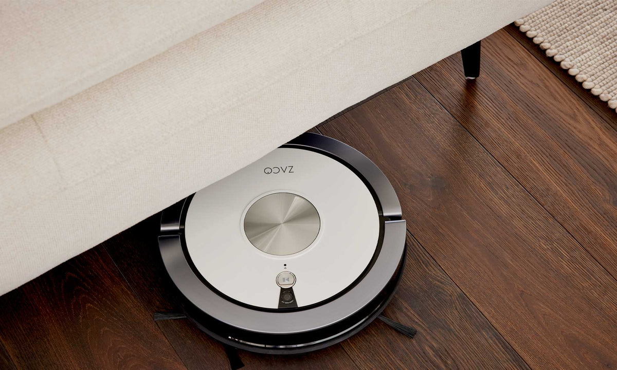 Ein runder Staubsaugerroboter reinigt unter einem ZEHDENICK | IL Interliving Sofa der Serie 4306 Jubiläums-Edition auf dunklem Holz.