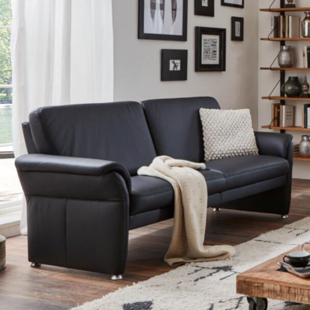 Das Sofa „Steinpol Polsteria Bornholm II“ mit weißem Kissen und cremefarbener Decke in einem gemütlichen, modernen Wohnzimmer.