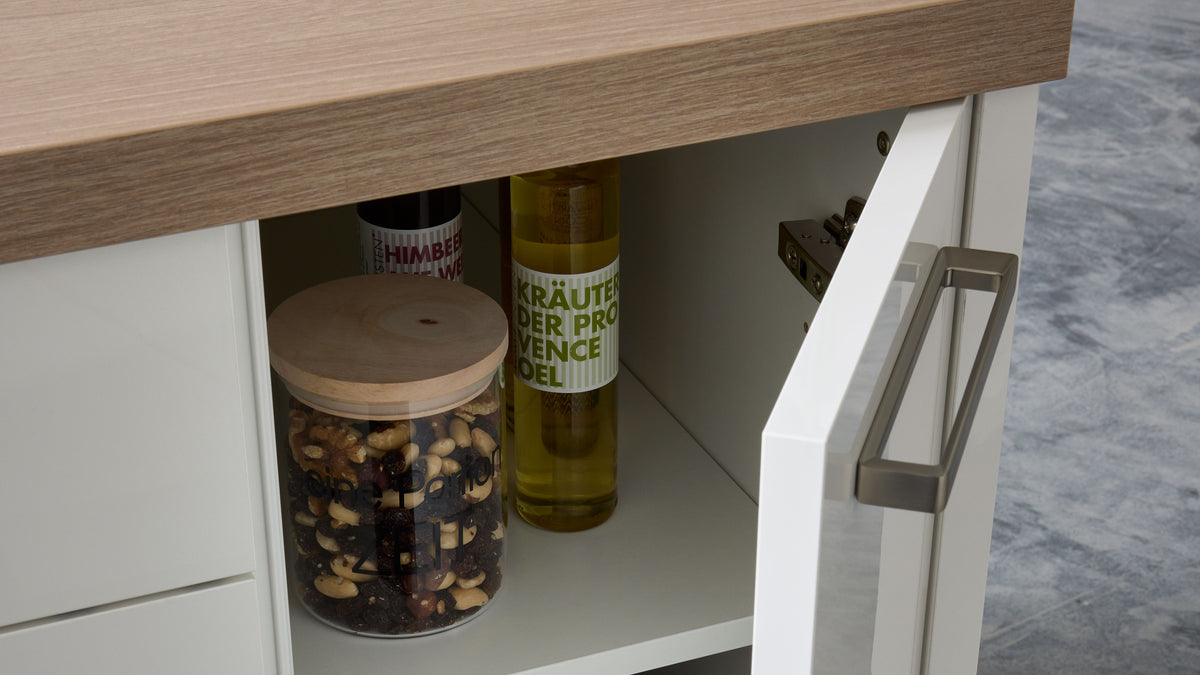 Offenes weißes Küchenfach unter Holzarbeitsplatte mit Glas voller Nüsse, einer hohen Ölflasche mit grünem Etikett und Türgriff.