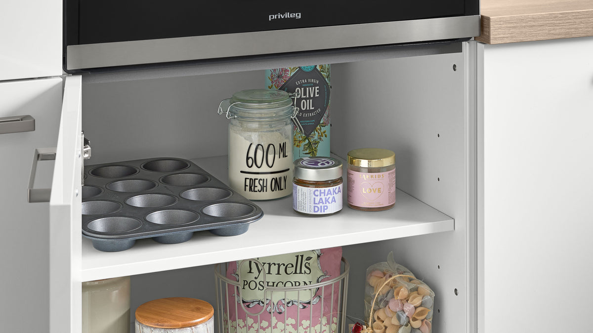 Offenes Küchenfach mit Metall-Muffinblech, Glasdose '600 ML FRESH ONLY', Olivenölkarton, kleine Gläser und Popcorntüte.