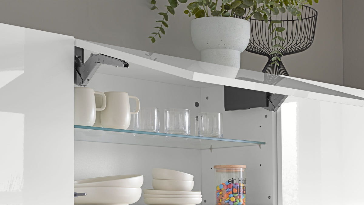 Weißer Hängeschrank mit geöffneter Klappe, Glasregal mit zwei Tassen, Gläsern, Schüsseln und Bonbonglas; oben Pflanze und Drahtkorb.