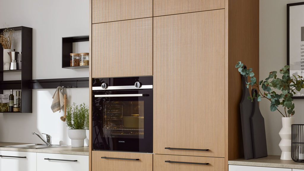 Hoher hellbrauner Küchenschrank mit Fronten und schwarzem Einbaubackofen, links Spüle und Regale, rechts Pflanze und Schneidebretter.