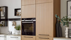 Hoher hellbrauner Küchenschrank mit Fronten und schwarzem Einbaubackofen, links Spüle und Regale, rechts Pflanze und Schneidebretter.