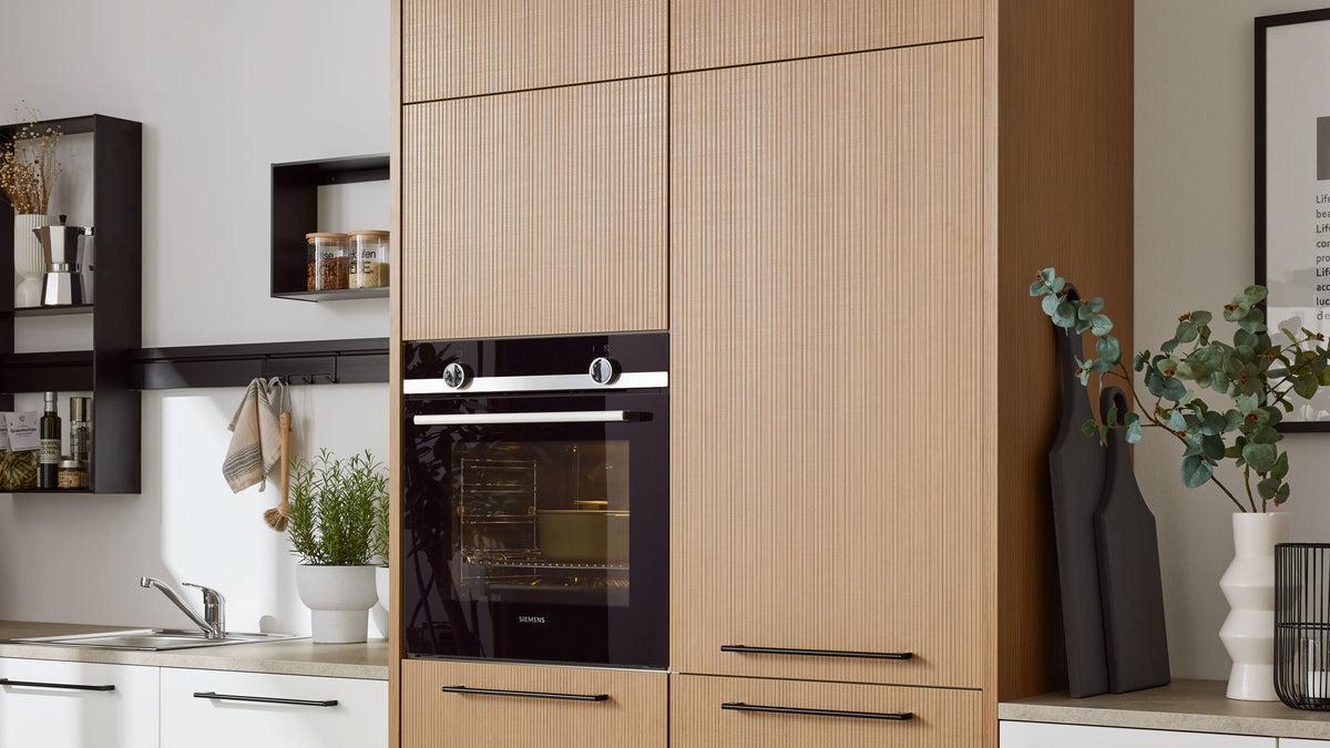 Hoher hellbrauner Küchenschrank mit Fronten und schwarzem Einbaubackofen, links Spüle und Regale, rechts Pflanze und Schneidebretter.