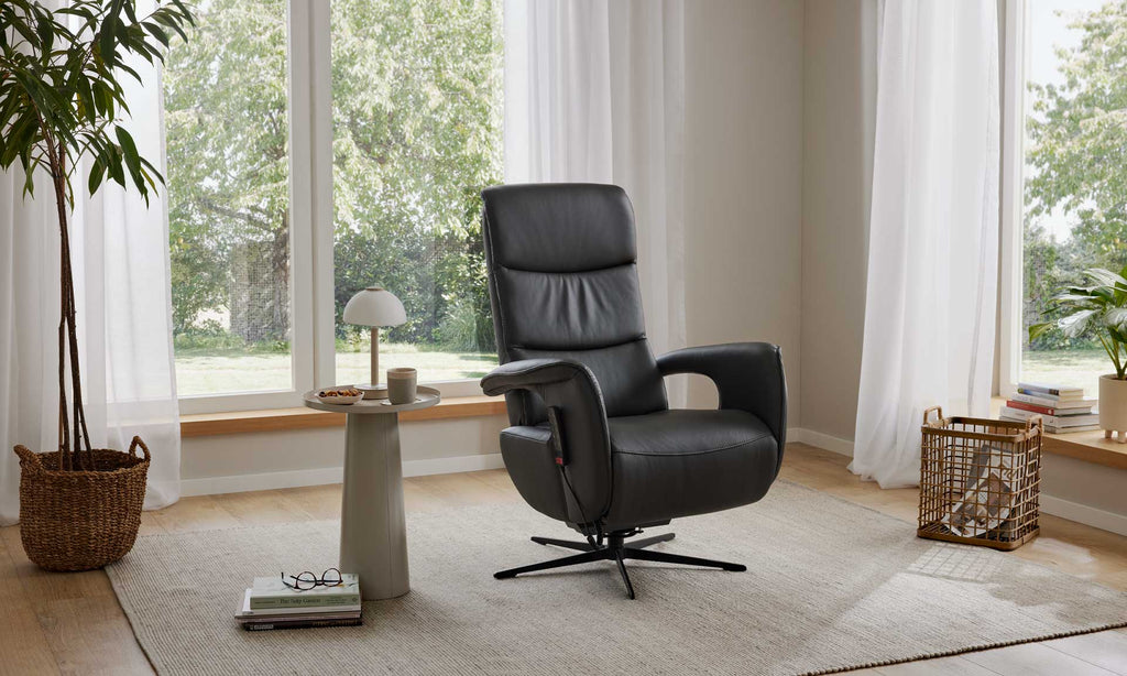 Ein HUKLA® | IL Relaxsessel aus der Interliving-Serie 4545 aus Glattleder steht auf einem beigen Teppich neben Grünpflanzen und großen Fenstern.