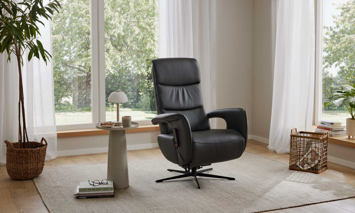 Ein HUKLA® | IL Relaxsessel aus der Interliving-Serie 4545 aus Glattleder steht auf einem beigen Teppich neben Grünpflanzen und großen Fenstern.
