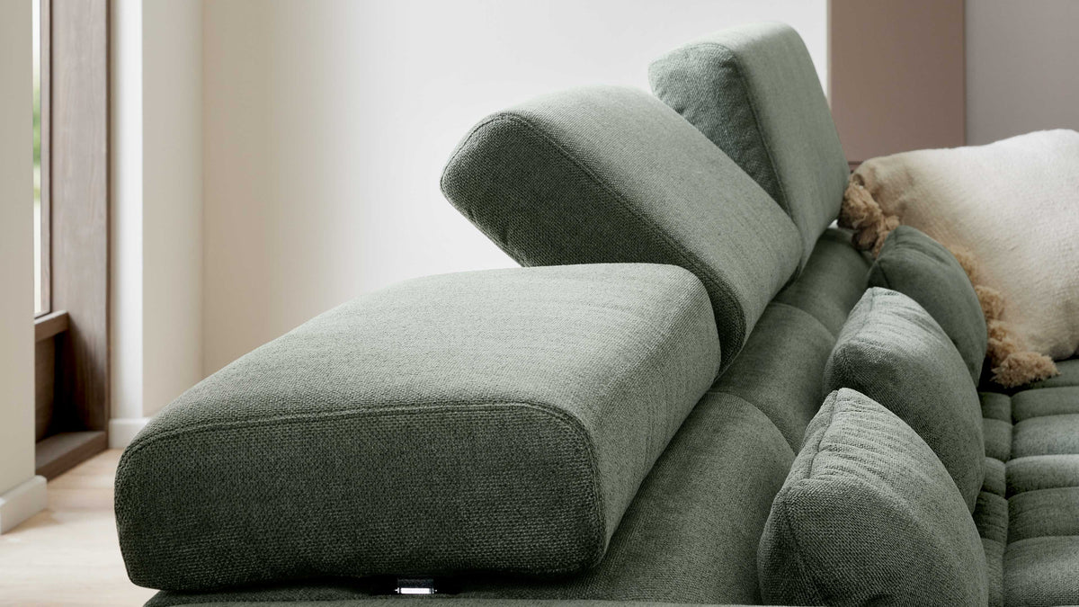 Grünes Stoffsofa mit verstellbarer Kopfstütze, mehreren gepolsterten Rückenkissen und sichtbarer Gewebestruktur, rechts helles Kissen.