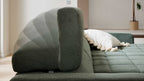 Grünes, stoffbezogenes Sofa mit mehrfach überlagerter verstellbarer Rückenlehne, im Hintergrund ein cremefarbenes Kissen mit Tasseln.