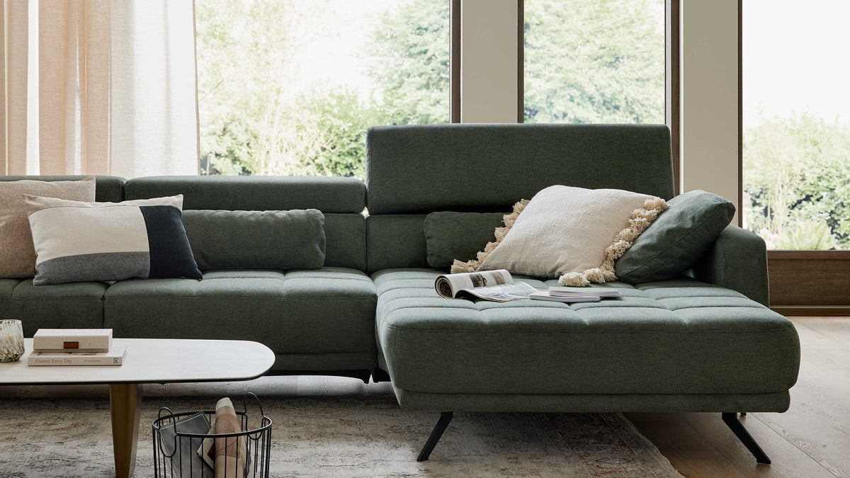 Grün-graue Eckcouch mit Recamiere, mehreren Zierkissen und aufgelegtem Magazin, davor niedriger Couchtisch.