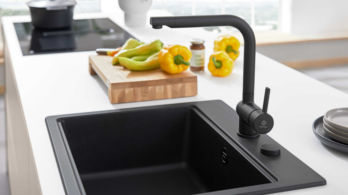 Schwarzer Wasserhahn und schwarze Einbauspüle auf weißer Arbeitsplatte, im Hintergrund Holz‑Schneidebrett mit gelben Paprika.