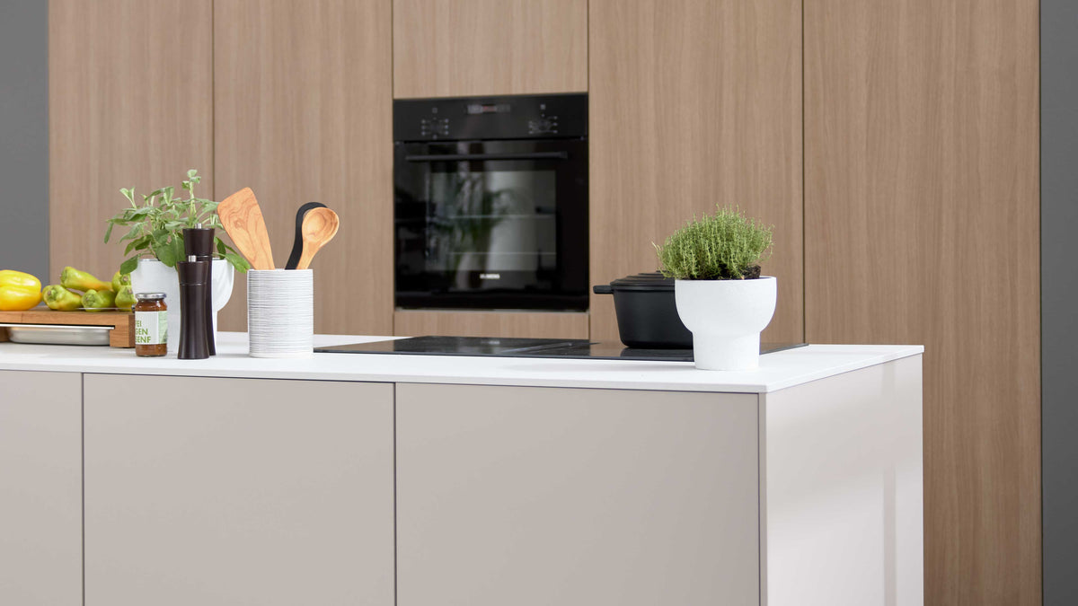 Kücheninsel mit Arbeitsplatte und Kochfeld, darauf Topf, Kräutertopf, Besteckbehälter und Gewürze; im Hintergrund Holzschränke mit Backofen.